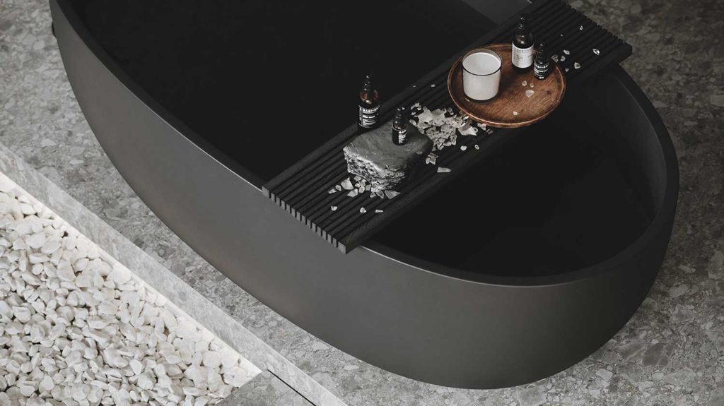 Une baignoire noire mate avec une planche de produits de soins sur une plateforme en terrazzo gris