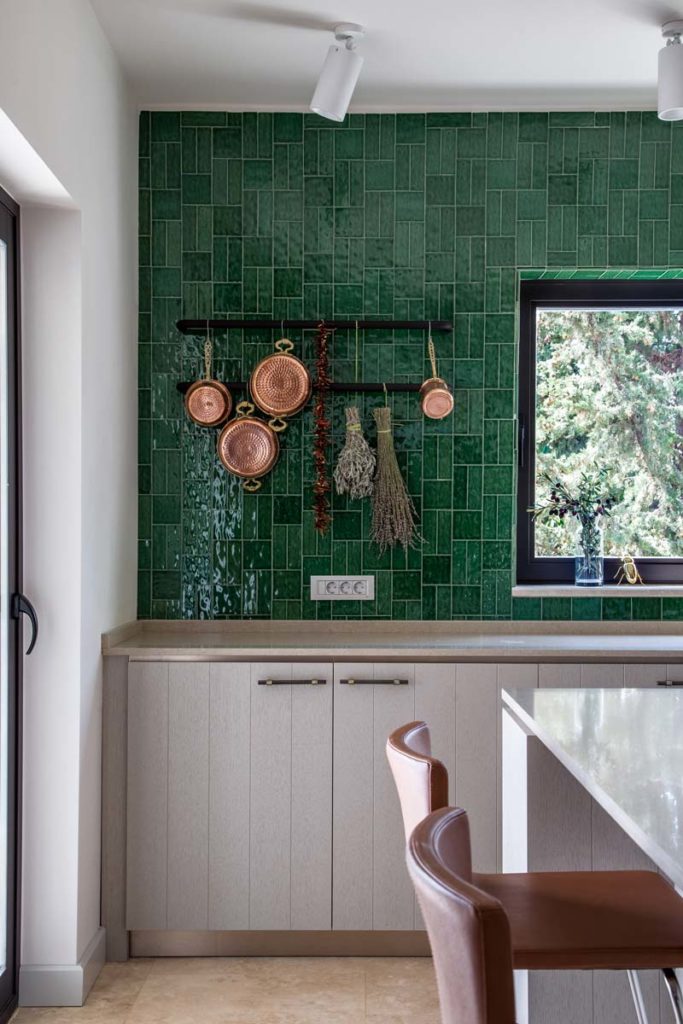 Un mur de cuisine en zellige vert avec des casseroles en cuivre et des meubles en bois blond