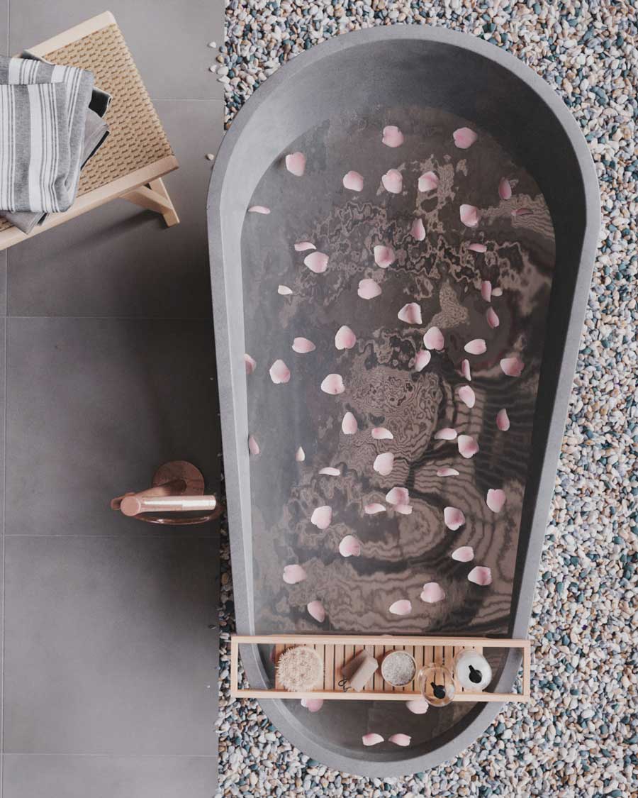 Une grande baignoire grise sur un sol en gravier, avec un panier en bois blond pour porter des produits de soin, et une robinetterie rose gold