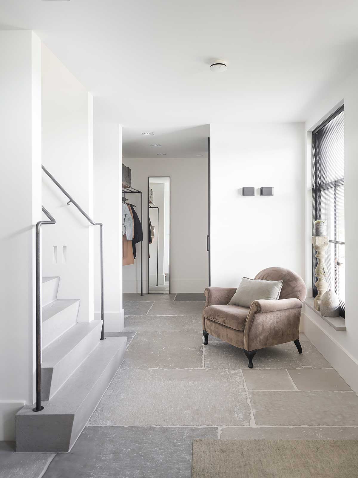 Un intérieur moderne blanc et gris avec des escaliers en béton, un sol en travertin et un fauteuil en velours