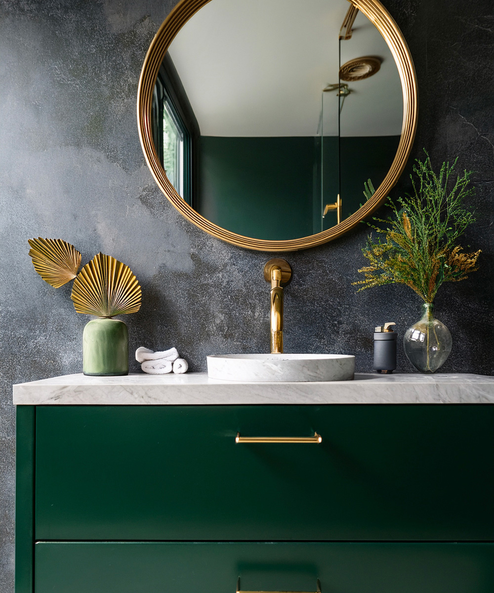 Salle de bain, avec miroir avec contour rond doré, robinetterie dorée, vasque en pierre, et tiroir vert et mur béton ciré en fond.